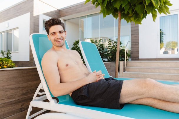 Foto de un feliz feliz joven positivo cerca del hotel al aire libre se encuentra en la silla de cubierta
. - Foto, imagen