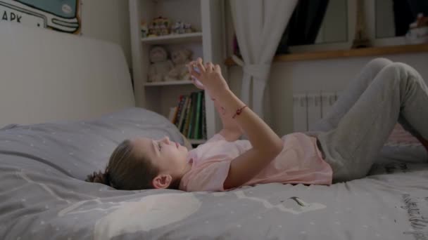 Niña linda juega en el teléfono inteligente acostado en la cama
 - Metraje, vídeo