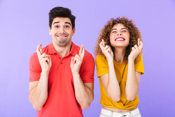 Portrait of caucasian people man and woman in basic clothing keeping fingers crossed and asking for good luck isolated over purple background - Foto, immagini