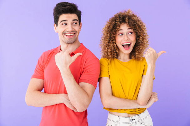 Portrait of happy caucasian people man and woman in basic clothing smiling and pointing fingers aside at copyspace isolated over violet background - Фото, изображение