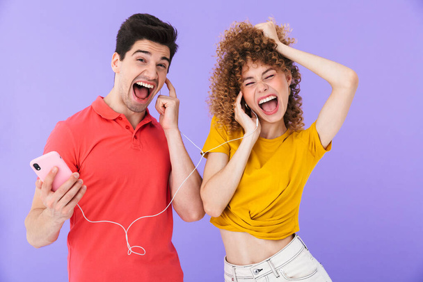 Portrait of happy caucasian people man and woman listening to music together with cellphone and earphones isolated over violet background - Photo, Image