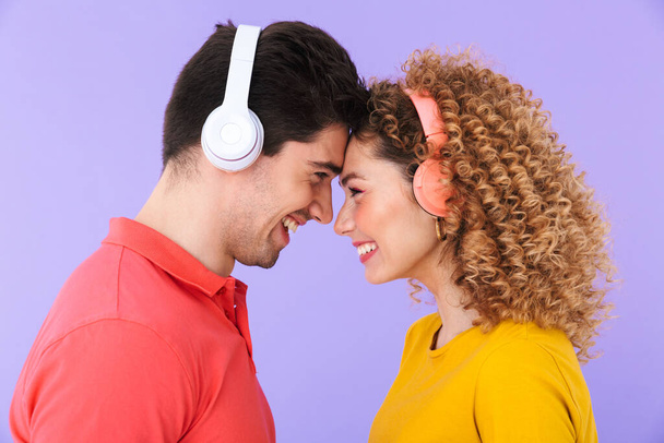 Portrait of beautiful couple smiling and listening to music with headphones while standing head to head isolated over violet background - Zdjęcie, obraz