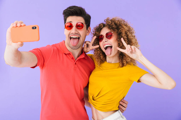 Portrait of excited couple smiling and showing peace sign while taking selfie photo on cellphone in trendy sunglasses isolated over violet background - Photo, image