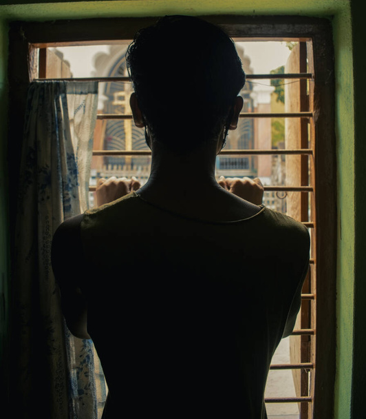 Rückseite Silhouettenfoto eines männlichen Typen, der aus dem Fenster nach draußen schaut und Fensterstangen hält - Foto, Bild