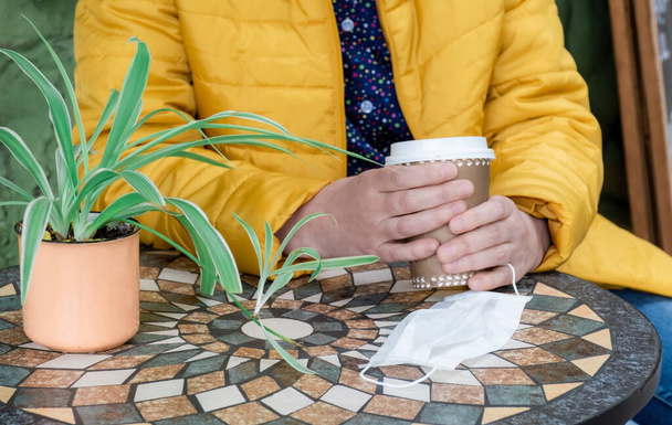 Manos femeninas sosteniendo taza de café y mascarilla médica. Mujer sentada fuera en la cafetería, concepto de café para llevar. - Foto, Imagen