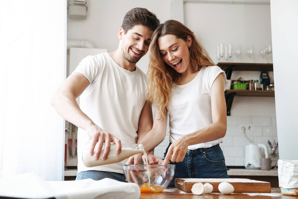 Image of positive young couple rejoicing while cooking breakfast together with milk and eggs in apartment - Foto, afbeelding