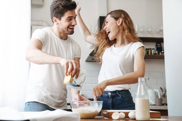 Image of positive couple rejoicing while cooking breakfast and mixing dough in kitchen at home - Fotó, kép