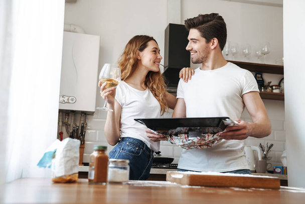 Image of joyous couple cooking together and holding baking pan with pastry in kitchen at home - Photo, Image