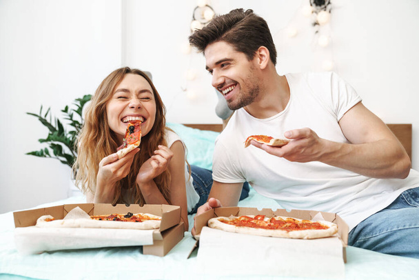 Image of caucasian happy couple man and woman lying on bed at home and eating pizza - 写真・画像