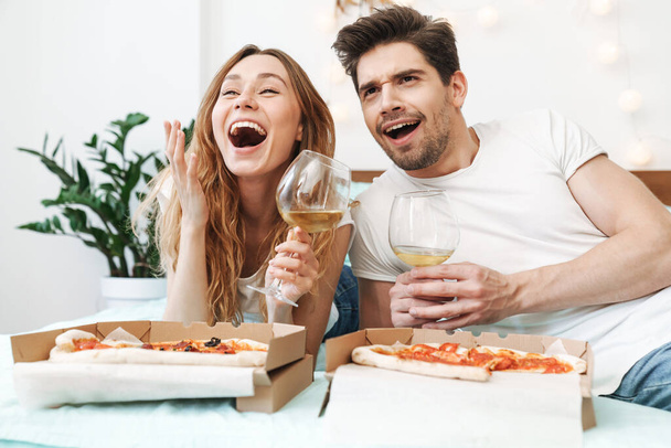 Image of happy couple man and woman drinking wine and eating pizza while lying on bed at home - Foto, Imagen