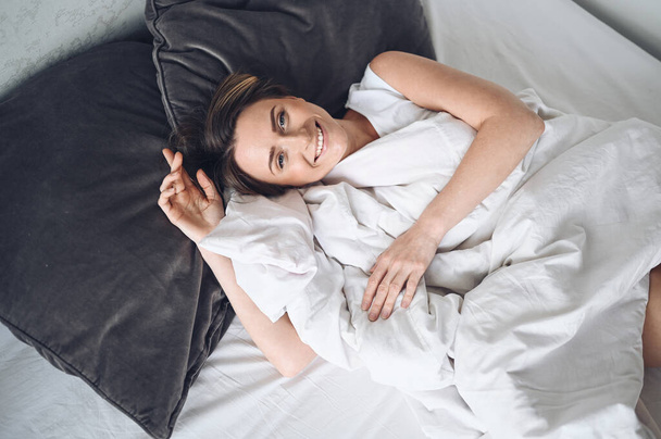 Ελκυστική χαμογελαστή νεαρή γυναίκα που τεντώνεται στο κρεβάτι ξυπνώντας μόνη χαρούμενη έννοια, ξύπνια μετά από υγιή ύπνο σε άνετο άνετο κρεβάτι και στρώμα απολαύστε καλημέρα - Φωτογραφία, εικόνα