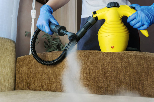 cleaning sofa with steam generator, hands in blue glove hold steam genetator and hose with brush, which produces steam for disinfection from microbes, dirt, allergy and viruses, home cleaning concept - Foto, Imagem