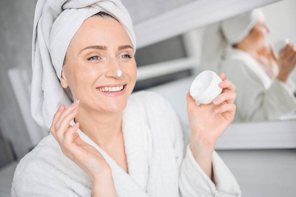 Cosmetology, skin care, face treatment, spa, natural beauty concept. Beautiful smiling woman at home in bathrobe with a towel applying face moisturizing cream from white jar. Beauty routine  - Fotografie, Obrázek