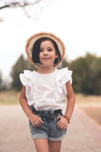 Smiling trendy kid girl 4-5 year old wearing straw hat and summer white top with denim shorts outdoors over nature background closeup.  - Fotografie, Obrázek