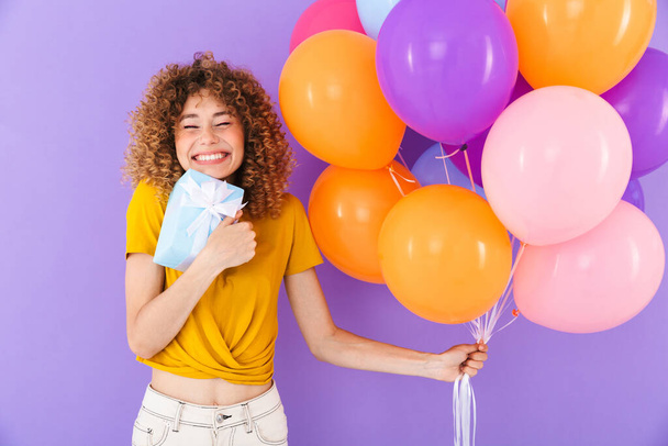 Image of happy young woman celebrating birthday with multicolored air balloons and present box isolated over violet background - Фото, зображення