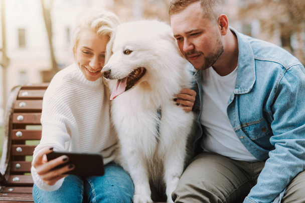 Smiling woman and man sitting on an alley bench with white fluffy puppy samoyed breed, making selfie by smartphone - Zdjęcie, obraz