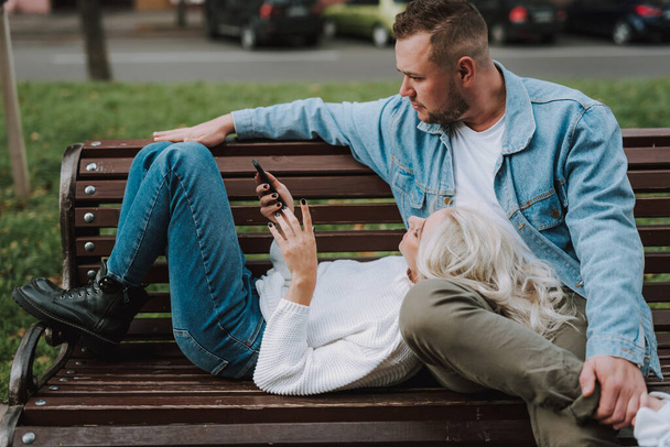 Happy blonde lady watching video on mobile phone while having rest with her boyfriend on an alley bench. Romantic date - Fotó, kép