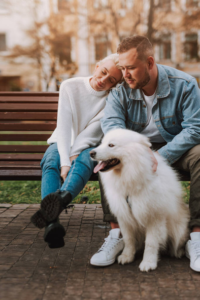 Young happy woman and man are enjoying their date while sitting on an alley bench with white fluffy puppy - Photo, Image