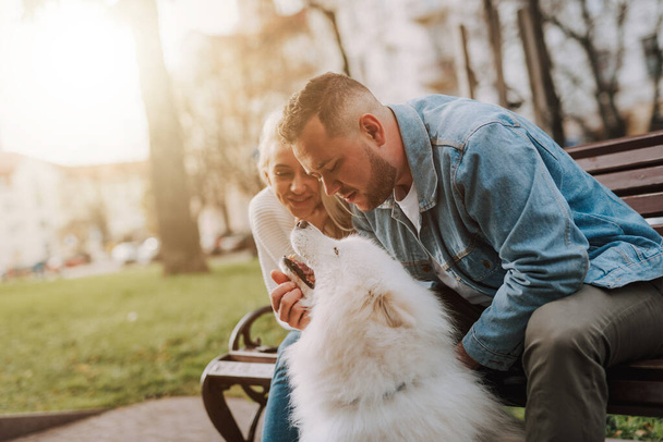 Молода щаслива усміхнена жінка і чоловік погладжують свого білого пухнастого собаку, сидячи на лавці
 - Фото, зображення