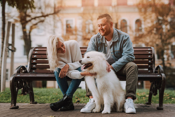 Young pretty female and male are enjoying date while sitting on an alley bench with white furry dog samoyed breed - Photo, Image