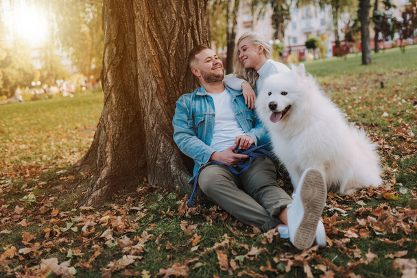 Happy pretty lady and her boyfriend posing with white furry playful puppy samoyed breed and sitting near big tree in autumn park - Фото, изображение