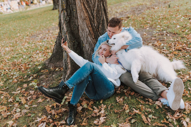 Smiling young couple with dog sitting in the park while making photo on smartphone - Foto, Imagen