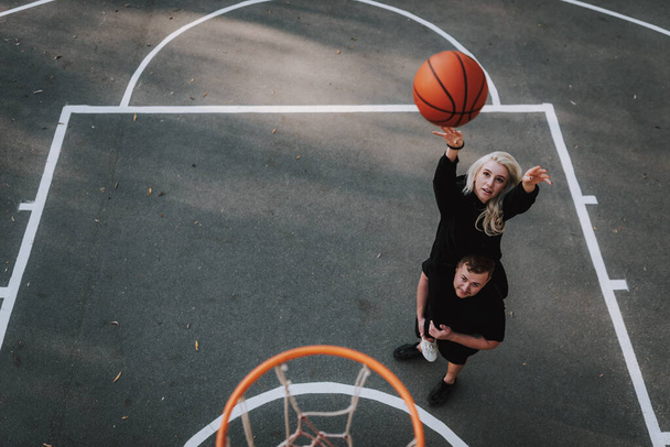 Top uitzicht van aantrekkelijke dame zitten op vriendje schouders tijdens het spelen van basketbal buiten - Foto, afbeelding