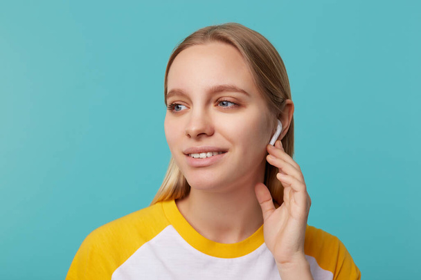Portrait of young attractive blue-eyed blonde long haired lady with earphones looking thoughtfully aside and smiling gently, isolated over blue background - Fotografie, Obrázek