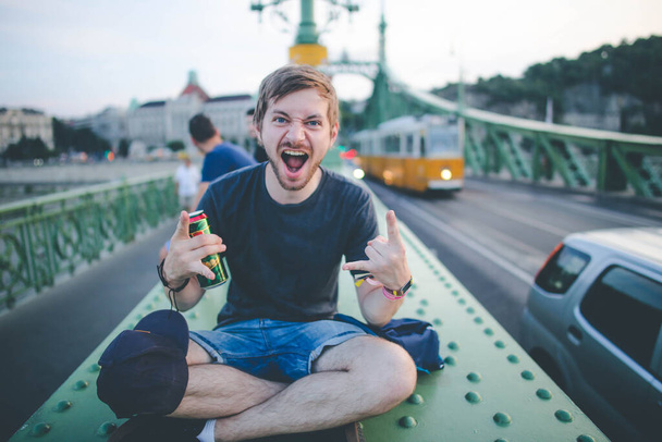 Νεαρός χαρούμενος γενειοφόρος άνδρας κάθεται στη γέφυρα στη Βουδαπέστη και κρατώντας μια κονσέρβα μπύρας, κοιτάζοντας χαρούμενα στην κάμερα - Φωτογραφία, εικόνα