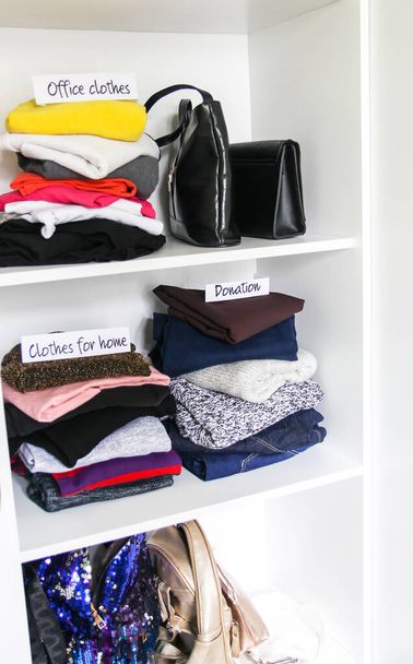 Kleidung und Accessoires sortieren im heimischen Kleiderschrank auf weißem Regalhintergrund. Bürokleidung, Kleidung für zu Hause, Scheine aus Spendenpapier - Foto, Bild