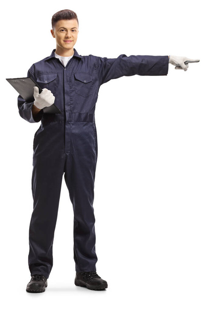 Full length portrait of a mechanic in a uniform holding a clipboard and pointing to the side isolated on white background - 写真・画像