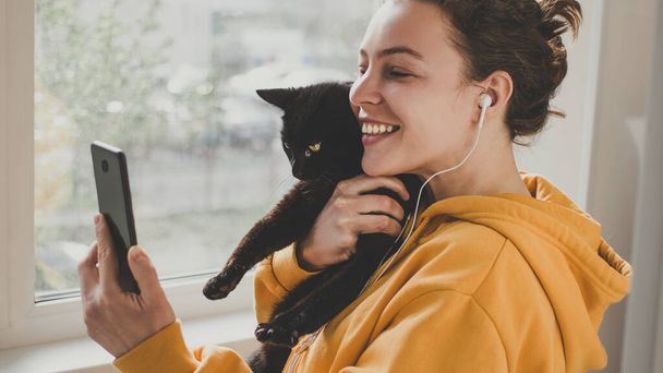 ヘッドフォンを装着した陽気な若い女性は、友人や親にこんにちはジェスチャー、ビデオ通話のためのスマートフォンを使用して黒いペット猫を保持しています。Happy smiling hipster girl making facetime zoom video calling - 写真・画像