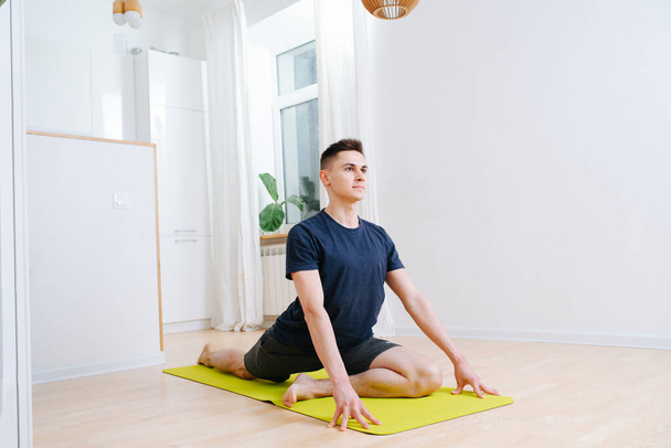 Tall man doing yoga on a mat in a kitchen at home during isolation. Lifting upper body with hands, with legs on a mat. Leg behind is straight, leg in front is folded. - Foto, Imagen