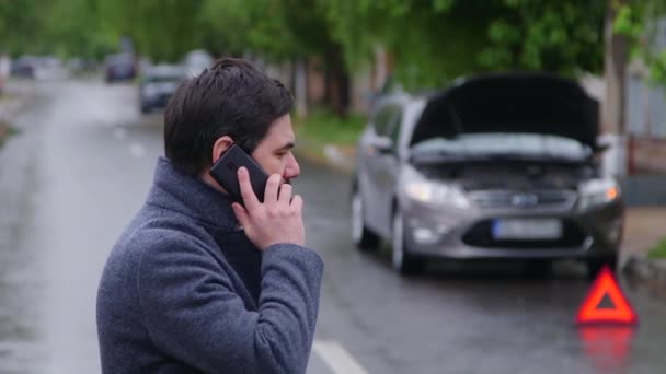 Nuori järkyttynyt liikemies seisoo rikki auton etsimässä korjauspalvelua älypuhelimella huolestuttava stressiä. Auto-onnettomuus. Ongelmia auton kanssa. - Materiaali, video