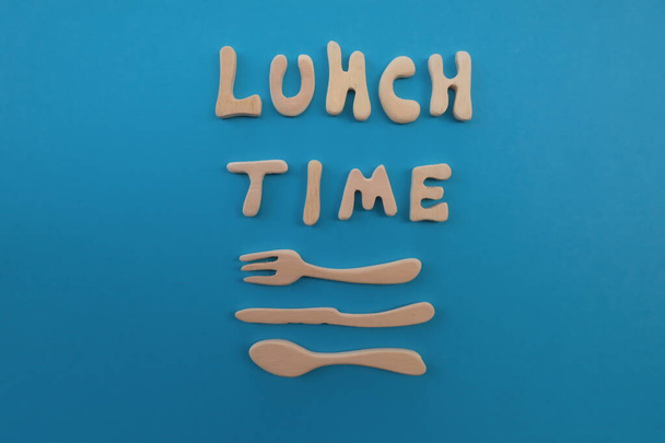 Texto a la hora del almuerzo compuesto con letras artísticas de madera con tenedor de madera, cuchillo y cuchara sobre fondo azul
 - Foto, imagen