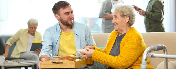Молодой человек подает ужин для пожилой женщины в гериатрическом хосписе, дизайн баннеров. Уход за пожилыми людьми
 - Фото, изображение