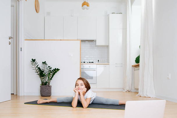 Little school age girl doing gymnastics on a yoga mat at home in isolation. Looking to laptop for instruction. She's sitting in splits, leaning chin on her hands. - Foto, immagini