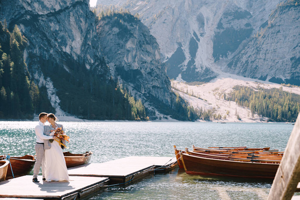Gelin ve damat İtalya 'daki Lago di Braies' deki ahşap bir tekne iskelesinde yürüyorlar. Avrupa 'da düğün, Braies Gölü' nde. Yeni evliler kayalık dağların arka planında yürür, öpüşür, sarılır.. - Fotoğraf, Görsel
