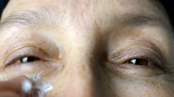 Зрелая взрослая женщина, кавказская национальность мажет увлажняющие косметические кремовые морщины на лице пальцами, смотрит в камеру. Крупный план
. - Кадры, видео