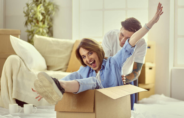 Couple having fun laughing moving into new home, young woman riding sitting in cardboard box while man pushing it - Foto, Bild