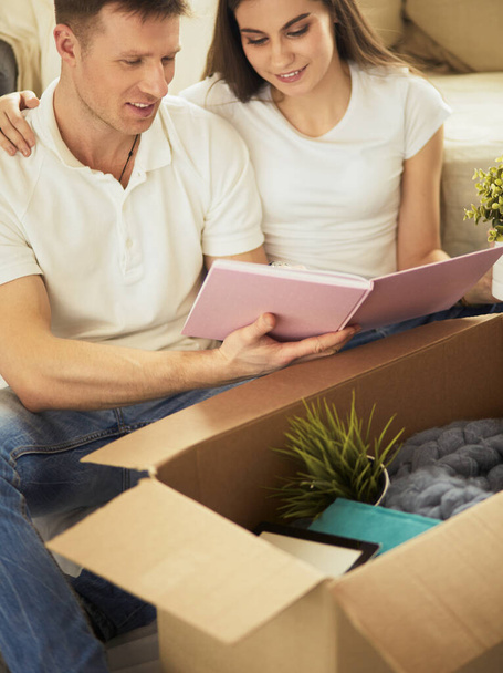 Cute couple unpacking cardboard boxes in their new home, sitting on the floor and looking at a family album - Φωτογραφία, εικόνα