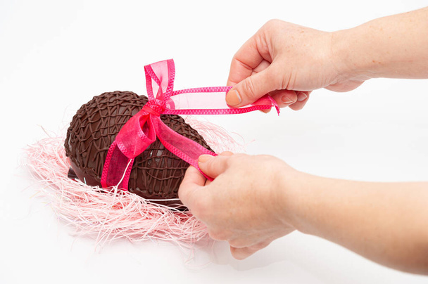 手作りのチョコレートの卵から赤い弓を開ける手。イースター、母の日、バレンタインデーのお祝い。白地に隔離された. - 写真・画像