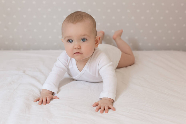 A little baby is smiling lying on the bed on a white sheet in the parents bedroom. A white jumpsuit in newborn. Cute baby lying on the bed. Closeup portrait of newborn baby looking at camera - Photo, Image