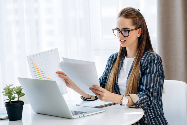 Δουλειά στο σπίτι. Κομψά ντυμένη κοπέλα με γυαλιά εργάζεται σε ένα φορητό υπολογιστή και με έγγραφα, ενώ κάθεται στο χώρο εργασίας της στο σπίτι - Φωτογραφία, εικόνα