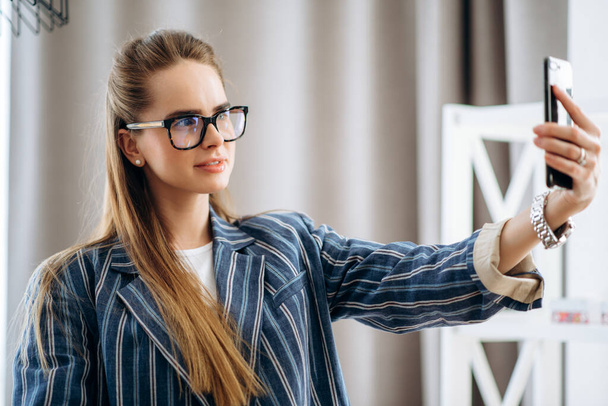 Confident girl in stylish wear and glasses smiling and makes selfie on the phone, indoors - Foto, Imagem