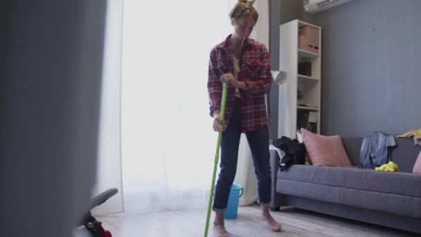 Happy young woman cute housewife is washing floor with mop and dancing at home - Filmmaterial, Video