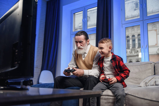 Netter kleiner Junge mit Großvater sitzt auf dem Sofa und spielt Videospiel mit Spielkissen - Foto, Bild