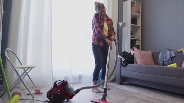 Young woman talking on the phone while cleaning the floor with a vacuum cleaner at home. - Πλάνα, βίντεο