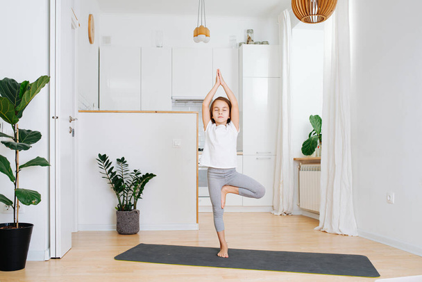 Little girl doing yoga on a yoga mat at home, standing on one leg with hands joined above her head. - Valokuva, kuva