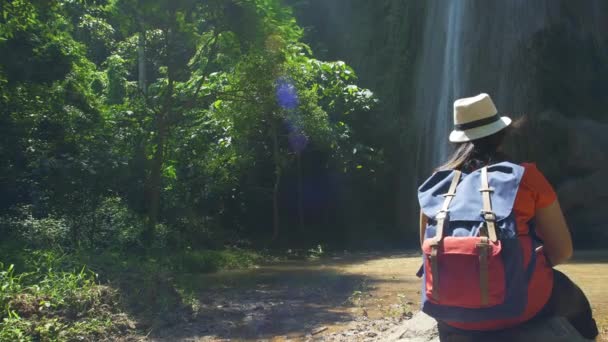 Widok z tyłu Backpacker Azjatki w lesie i relaksujące, korzystających siedzieć patrząc w wodospadzie. Turyści piesze wycieczki w lesie przygoda podróży Azja Tajlandia - Materiał filmowy, wideo